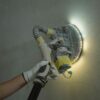 Close-up of a worker sanding a wall with a power tool under bright lighting.
