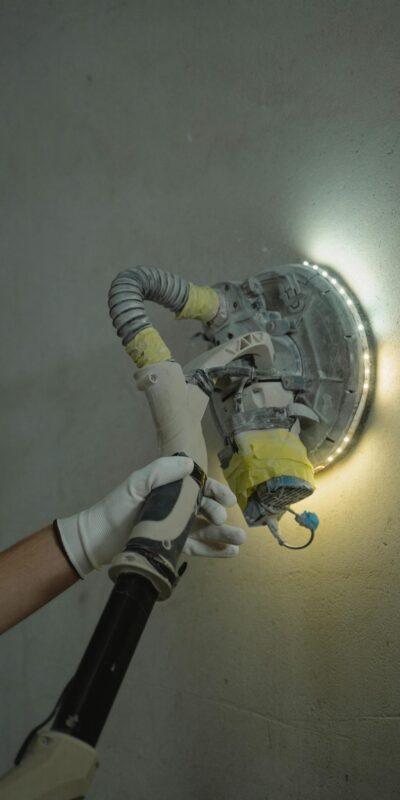 Close-up of a worker sanding a wall with a power tool under bright lighting.